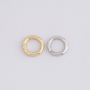 Könnte beinhalten: Zwei runde Metallverschlüsse, einer goldfarben und einer silberfarben, vor weißem Hintergrund. Die kreisförmigen Verschlüsse mit Scharnieröffnung sind für Schmuck oder Bastelprojekte geeignet. Der goldfarbene Verschluss hat eine glänzende Oberfläche.