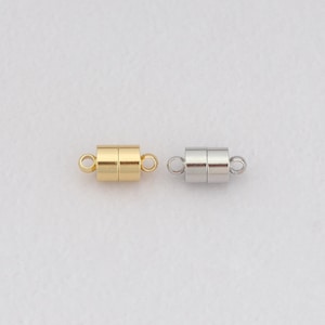 May include: Two magnetic jewelry clasps. One is gold-colored, the other is silver-colored. Each clasp is cylindrical with a small loop on each end for attaching to jewelry. The clasps are shown against a white background.