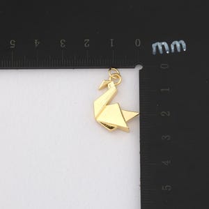 K&ouml;nnte beinhalten: Ein goldfarbener Origami-Schwan-Anh&auml;nger. Der Anh&auml;nger besteht aus gefaltetem Metall und hat oben eine Schlaufe zum Befestigen an einer Kette. Der Schwan befindet sich auf einer wei&szlig;en Oberfl&auml;che neben einem schwarzen Lineal.