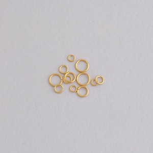 May include: A selection of small, gold-coloured jump rings of various sizes, scattered on a light grey surface. The rings are circular and appear to be made of metal, with a shiny finish. The sizes range from very small to medium, suitable for jewellery making or crafts.