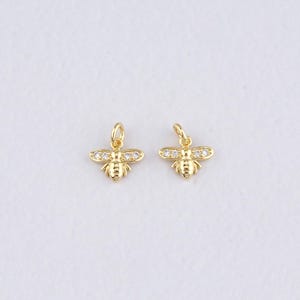 May include: Two gold-colored bee charms with small, clear crystals on the wings. Each charm has a loop at the top for attaching to a chain or bracelet. The bees have detailed bodies and are set against a white background.