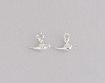 Puede incluir: Dos colgantes plateados con forma de lámpara de genio. Cada colgante presenta un diseño de lámpara clásico con un pico curvo, un asa y un pequeño lazo para sujetarlo a una cadena o pulsera. Sobre un fondo gris claro.