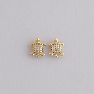 May include: A pair of gold-coloured turtle-shaped earrings. Each earring has a textured shell design with small, clear stones. The turtles have small legs and a head. The earrings are set against a plain, light background.