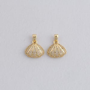 Könnte beinhalten: Zwei goldfarbene Muschel-Anhänger mit einer glitzernden, strukturierten Oberfläche. Jeder Anhänger hat oben eine kleine Schlaufe zum Befestigen an einer Kette oder einem Ohrringhaken. Die Anhänger sind vor einem schlichten weißen Hintergrund platziert.