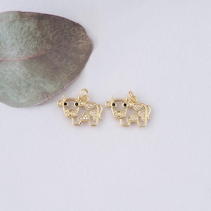 May include: Two gold-colored cow charms with black eyes and clear crystal accents. The charms are in a standing position, with a loop at the top for hanging. The charms are likely for jewelry making or crafting.