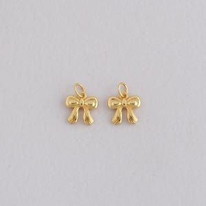 May include: Two gold-colored bow charms with a loop at the top for jewelry making. The bows have a detailed, textured design. These charms are suitable for earrings, necklaces, or bracelets.