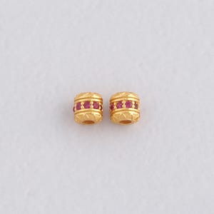 Op de afbeelding: Twee goudkleurige, cilindrische kralen met een herhalend patroon van kleine, rode edelstenen. De kralen hebben een getextureerd oppervlak en worden tegen een witte achtergrond getoond. De kralen zijn waarschijnlijk voor het maken van sieraden.