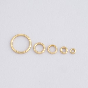 Pode incluir: Uma coleção de cinco anéis de metal dourado de vários tamanhos. Os anéis estão dispostos em uma fileira, com o anel maior à esquerda e o menor à direita. Os anéis têm um acabamento liso e polido.