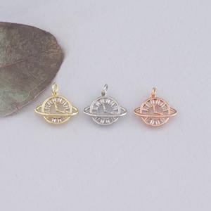 May include: Three gold, silver, and rose gold charm pendants in the shape of a clock with a ring around it, resembling a planet. The charms are on a white background.