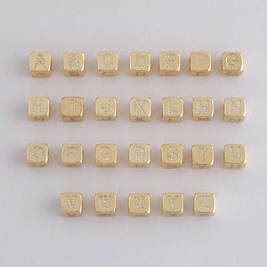 Puede incluir: Cuentas doradas con letras del alfabeto de la A a la Z, dispuestas en filas. Las letras están incrustadas con pequeños cristales transparentes. Las cuentas cuadradas tienen un brillo metálico, ideales para la creación de joyas.