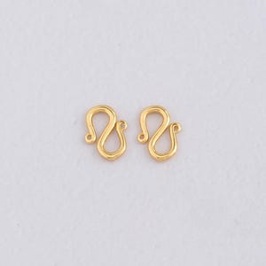 Puede incluir: Dos cierres de joyería dorados en forma de S. Cada cierre tiene un diseño curvo con un pequeño bucle en cada extremo. Los cierres se muestran sobre un fondo blanco, mostrando sus intrincados detalles.