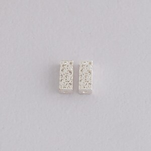 Puede incluir: Dos pendientes rectangulares plateados con un diseño calado de filigrana. Los pendientes tienen forma rectangular con un patrón decorativo de líneas y curvas arremolinadas. Los pendientes se muestran sobre un fondo blanco.