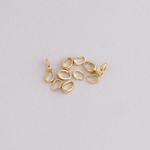 May include: A selection of small, gold-coloured jewellery findings, including oval-shaped connectors and a few clasp-style pieces. The findings are scattered on a white surface, with soft, diffused lighting.