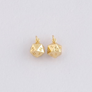 May include: Two gold-coloured, polyhedral dice charms, each with numbers on its faces. The dice are suspended from small loops, indicating they are for jewellery or crafts. The background is a plain, light colour.