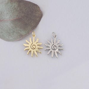 Pode incluir: Dois pingentes em forma de sol. Um é dourado, o outro prateado. Cada pingente tem um centro em espiral e raios radiantes. Os pingentes são pequenos, adequados para fazer joias ou como amuletos.