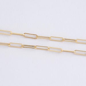 May include: Close-up of two gold-coloured paperclip chain necklaces. The chains are made of rectangular links connected by small, thin bars. The background is a plain white surface, highlighting the jewellery's design and colour.