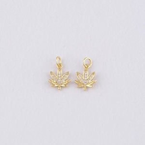 Puede incluir: Dos colgantes dorados con forma de hoja y detalles de cristal transparente. Cada colgante tiene un pequeño lazo en la parte superior para sujetarlo a una cadena u otra joya. Los colgantes están sobre un fondo blanco.
