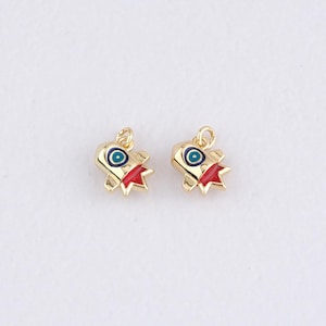 May include: Two gold-colored charms, each shaped like a stylized rocket ship. The rockets have red fins, blue and white eye-like details, and a loop for attaching to a chain or bracelet. The charms are set against a white background.