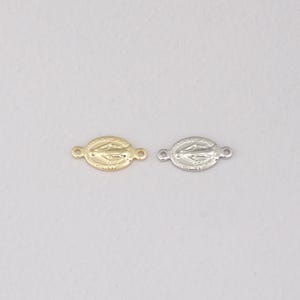 May include: Two oval connectors, one gold-toned and one silver-toned, featuring a religious design. Each connector has a loop at either end for attaching to other jewellery components. The connectors are displayed on a white background.