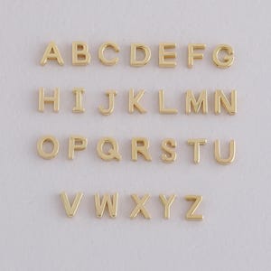 以下が含まれることがあります： 金色の金属製アルファベット文字、AからZまで、行に配置されています。各文字は、ソリッドなブロックデザインです。文字は均等に配置され、明るい背景に表示されており、ジュエリー作りやクラフトに適しています。