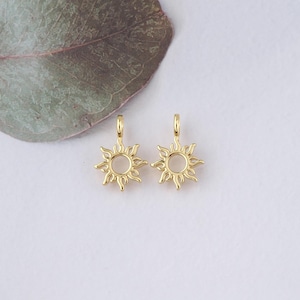 May include: Two gold-colored sun charm pendants with a circular opening in the center. The charms are designed with a detailed sun shape with rays extending outward.