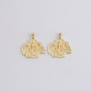 May include: Two gold-colored pendants with a textured, organic design. Each pendant has a small loop at the top for a chain. The pendants have a detailed, irregular shape, and are displayed against a plain white background.
