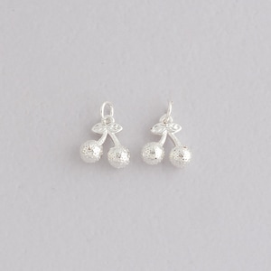 Peut inclure: Deux breloques argentées en forme de cerises. Chaque breloque présente deux sphères texturées représentant les cerises, reliées par une tige et des feuilles. Les breloques ont une petite boucle en haut pour la fixation.