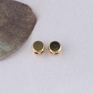 Puede incluir: Dos cuentas de metal doradas con dos agujeros cada una. Las cuentas son redondas y tienen una superficie lisa.