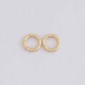 May include: Two gold-colored, round jewelry clasps with small, clear gemstones set around the outer edge. The clasps are open, revealing the clasp mechanism. The background is a plain, off-white color.