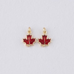 May include: Two gold-colored maple leaf charms with red enamel filling. Each charm has a small loop at the top for attaching to a necklace or bracelet. The charms are displayed on a white background.