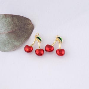 Puede incluir: Dos abalorios de cereza de color dorado con cerezas de esmalte rojo y hojas verdes.