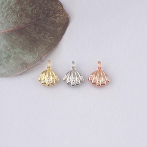 May include: Three seashell charms in gold, silver, and rose gold. Each charm features a shell design with small, clear gemstones. The charms are displayed on a white surface with a green leaf in the background.