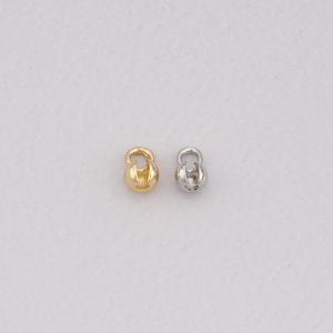 May include: Two small, dumbbell-shaped jewellery findings. One is gold-coloured, the other is silver-coloured. Both have a rounded body with a curved top, designed for jewellery making or crafting.