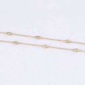 Puede incluir: Un delicado collar dorado con múltiples gemas ovaladas y transparentes. Las gemas están espaciadas uniformemente a lo largo de la fina cadena, creando un diseño minimalista y elegante. El collar se muestra sobre un fondo blanco liso.
