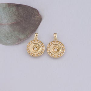 May include: Two gold-tone pendants with a circular design. Each pendant features a small, clear crystal in the center and a swirl design around it. The pendants are surrounded by a circle of smaller, clear crystals.