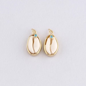 May include: Two gold-colored oval pendants, each featuring a cream-colored seashell and a small turquoise accent. The pendants have a loop at the top for hanging. The background is plain white.