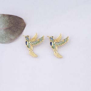 May include: A pair of gold hummingbird earrings with blue and green enamel accents. The earrings are detailed with small, sparkling stones.