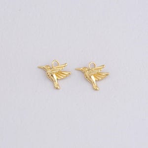 以下が含まれることがあります： 2つの金色のハチドリチャーム。各チャームは、翼を広げたハチドリの詳細なデザインで、長い嘴と、ジュエリーに取り付けるための上部の小さなループが特徴です。チャームは白い背景にあります。