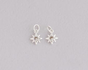 Puede incluir: Dos pequeños colgantes plateados con forma de estrella, cada uno con una gema transparente en el centro. Tienen un lazo en la parte superior para sujetarlos a joyas. Los colgantes se muestran sobre un fondo gris claro.