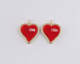 Könnte beinhalten: Zwei herzförmige Anhänger. Jeder Anhänger hat eine rote Mitte mit dem Wort "LOVE" in Weiß. Die Anhänger sind mit kleinen, klaren Steinen und einem goldfarbenen Metallrahmen umrandet. Sie haben oben eine kleine Schlaufe für eine Kette.