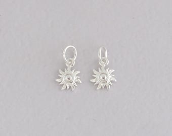 Puede incluir: Dos colgantes de sol plateados, cada uno con un círculo central y rayos puntiagudos. Tienen pequeños bucles para sujetarlos a joyas. Los colgantes se muestran sobre un fondo gris claro. Cada colgante mide aproximadamente 1 cm de diámetro.