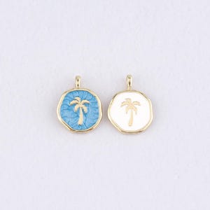 Puede incluir: Dos colgantes dorados con diseño de palmera. Uno tiene fondo azul claro, el otro es blanco. Ambos tienen contorno dorado y un pequeño lazo en la parte superior para sujetar a una cadena o pulsera.