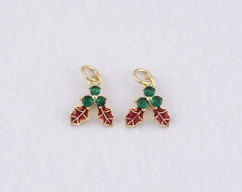 May include: Two gold-colored holly charms, each featuring three green gemstone berries and red enamel leaves. The charms have a small loop at the top for attaching to jewelry. The background is plain white.