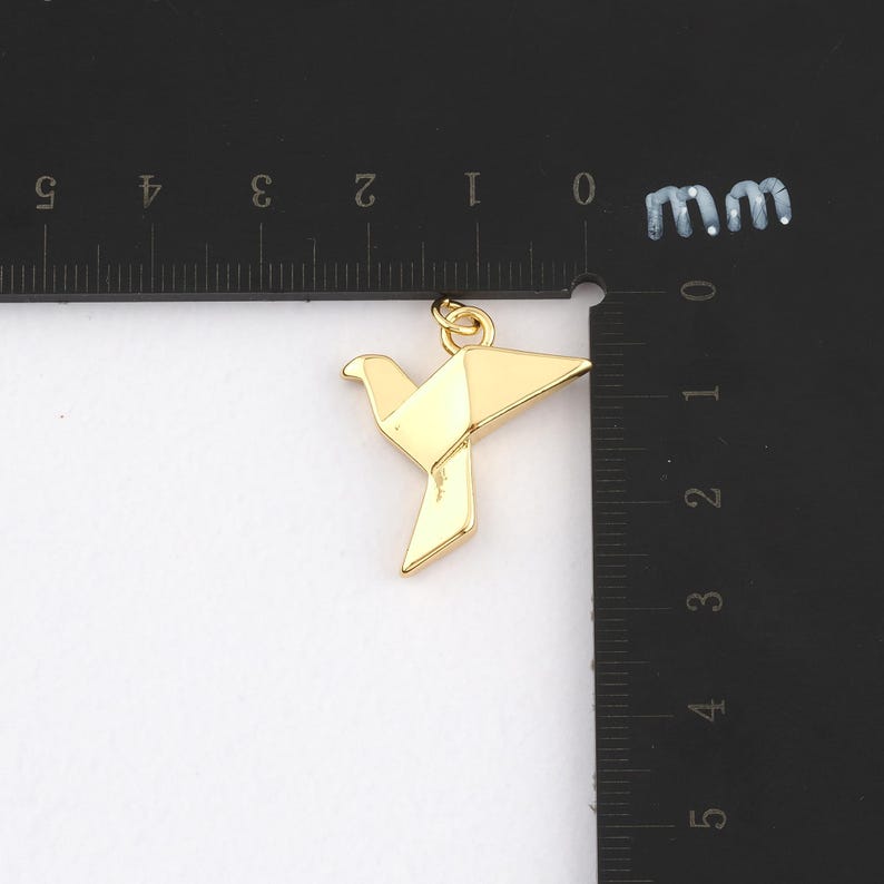 K&ouml;nnte beinhalten: Ein goldfarbener Origami-Vogel-Anh&auml;nger. Der Anh&auml;nger ist eine geometrische Darstellung eines Vogels mit gefalteten Fl&uuml;geln und einer kleinen Schlaufe oben f&uuml;r eine Kette. Der Anh&auml;nger ist etwa 2.5 cm lang.