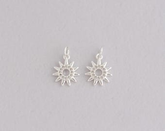 Puede incluir: Dos colgantes plateados con forma de sol, cada uno con un círculo central de piedras transparentes y rayos puntiagudos. Tienen pequeños bucles para sujetarlos a la joyería. Fondo gris claro.
