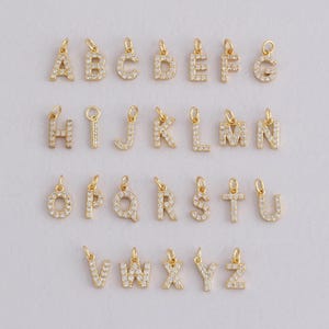 Puede incluir: Una colección de colgantes de alfabeto dorados, cada uno incrustado con pequeñas piedras preciosas transparentes. Se muestran las letras de la A a la Z, cada una con un pequeño lazo para sujetar. Los colgantes están dispuestos sobre un fondo claro.