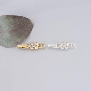 Könnte beinhalten: Zwei Schmuckverschlüsse, einer goldfarben und einer silberfarben, werden gezeigt. Jeder Verschluss hat ein Blumendesign, das mit kleinen, klaren Edelsteinen besetzt ist. Die Verschlüsse sind rechteckig mit einem sicheren Verschlussmechanismus.