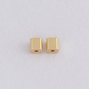 May include: Two small, gold-coloured, square beads with a hole drilled through the centre. The beads have a smooth, polished surface and are likely used in jewellery making or other craft projects. The beads are shown against a plain white background.