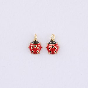 Peut inclure: Deux breloques dorées en forme de coccinelles, avec des corps rouges, des têtes noires et des détails dorés. Chaque breloque a une petite boucle pour suspendre. Les coccinelles ont des points noirs, sur fond blanc.
