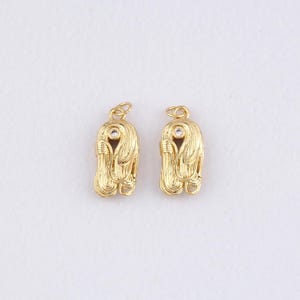 Könnte beinhalten: Zwei goldfarbene Anhänger mit strukturiertem Design. Jeder Anhänger hat einen zentralen klaren Stein und eine kleine Schlaufe oben zur Befestigung. Die Anhänger haben eine einzigartige, stilisierte Form.